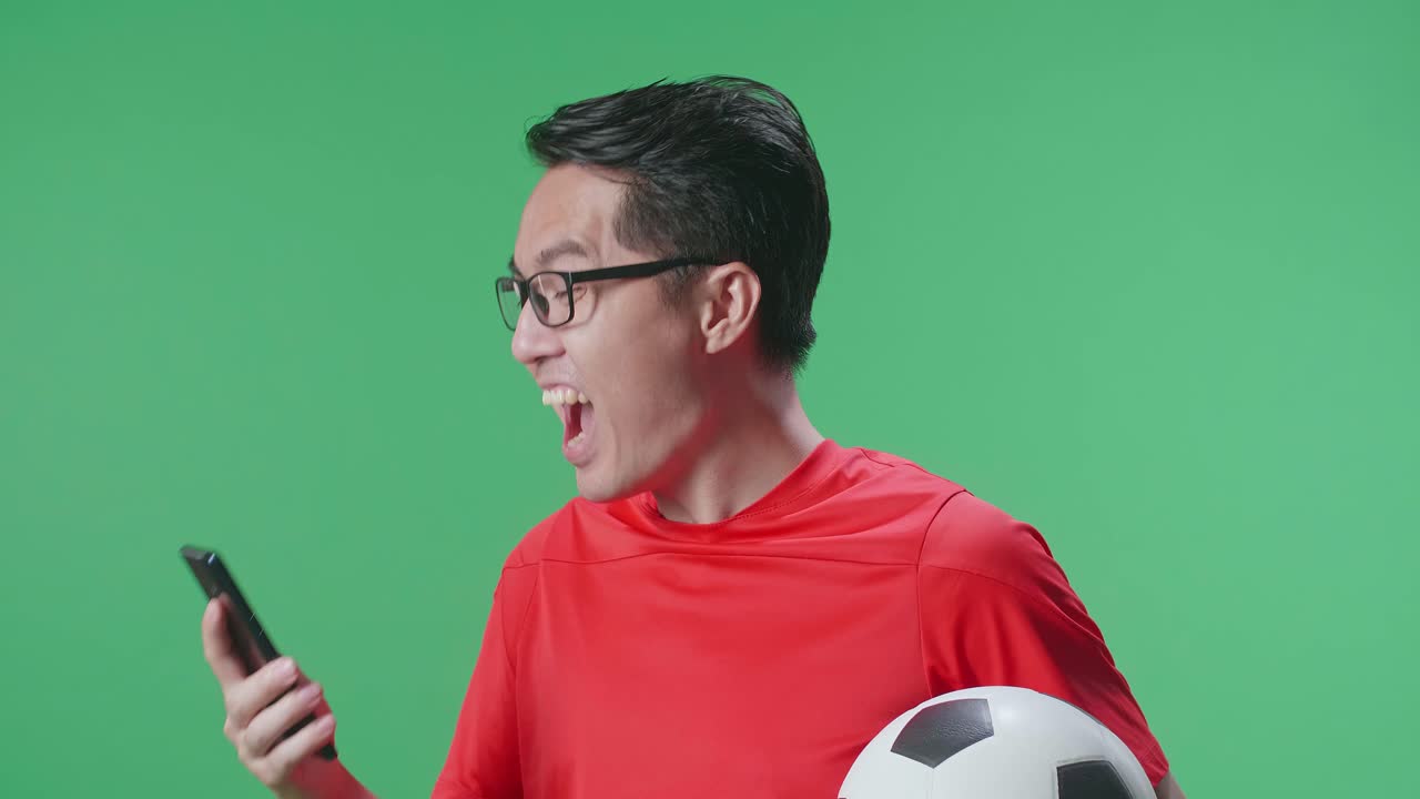 Close Up Side View Of Asian Man With A Ball Screaming Goal Celebrating While Cheering Soccer On Smartphone In Green Screen Background