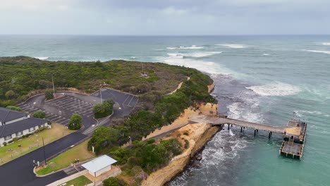 drone footage captures the scenic coastline and jetty at port campbell, australia, with lush greenery and ocean waves under cloudy skies