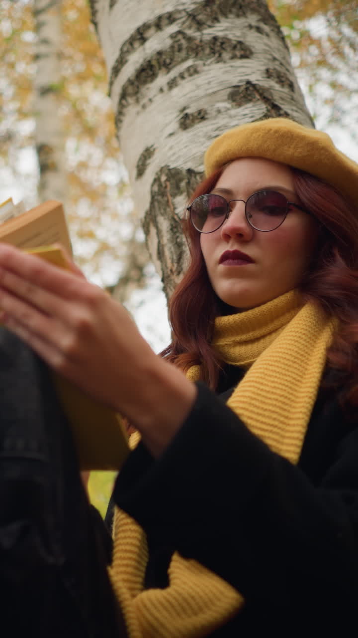 mujer con boina amarilla y bufanda sentada sola, apoyada en un árbol, abriendo cuidadosamente un libro cubierto de amarillo, rodeada de doradas hojas de otoño y abedules, disfruta de la lectura tranquila y la reflexión