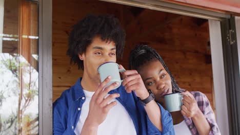 Happy-african-american-couple-looking-through-window-and-drinking-coffee-in-log-cabin,-slow-motion