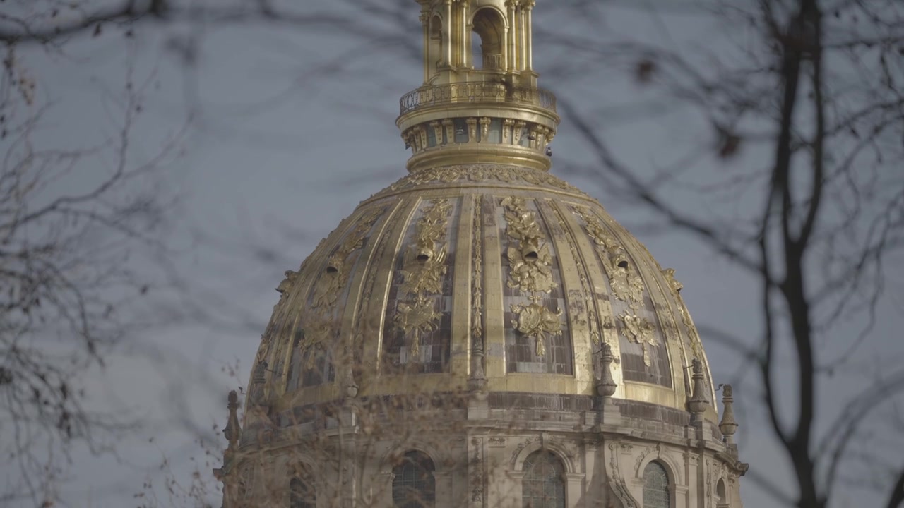 Una vista ravvicinata della cupola dorata e ornata de Les Invalides contro un cielo nuvoloso, incorniciata da rami d'albero.