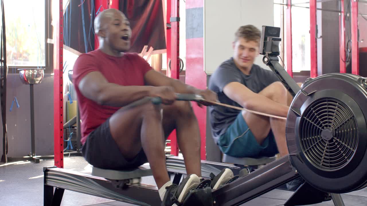 Diverse male duo in gymwear gripping handles rowing water-resistance rowers at gym for training