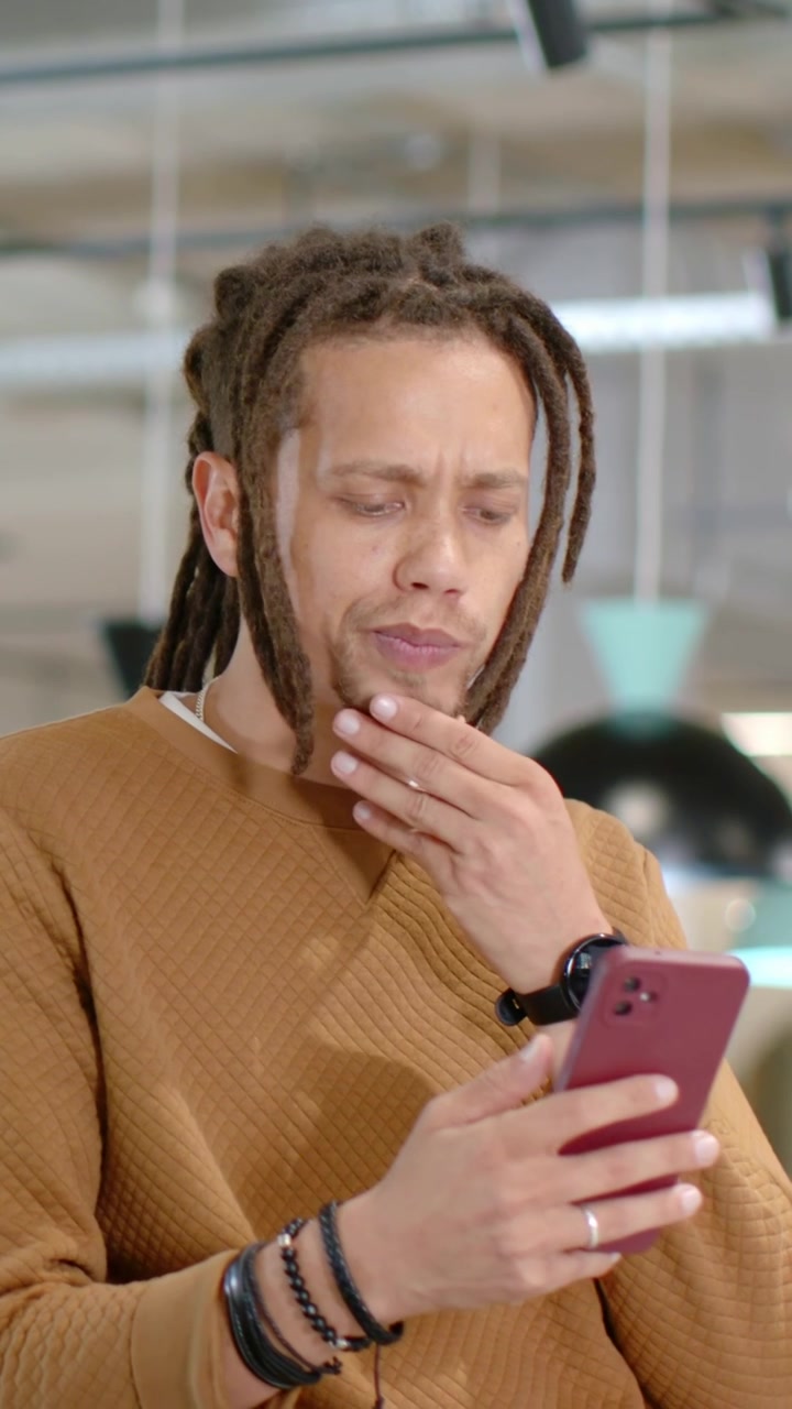 Vertical video: Reading text man with dreads in brown sweater tapping pink phone at cafe smiling