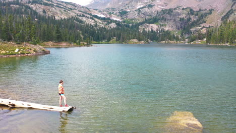 aerial drone flies over woman standing on the end of a fallen tree log in a clear blue water lake to ravleal mountain peak and a thick pine tree forest in nederland colorado rocky mountains