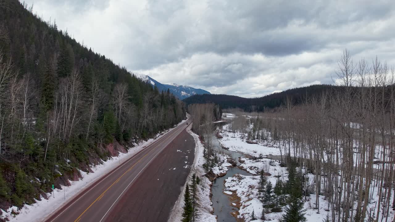 Ripresa aerea con drone di un'autostrada che si snoda attraverso una valle con sponde spolverate di neve, alberi sempreverdi e spogli e montagne sullo sfondo vicino a Whitefish, Montana