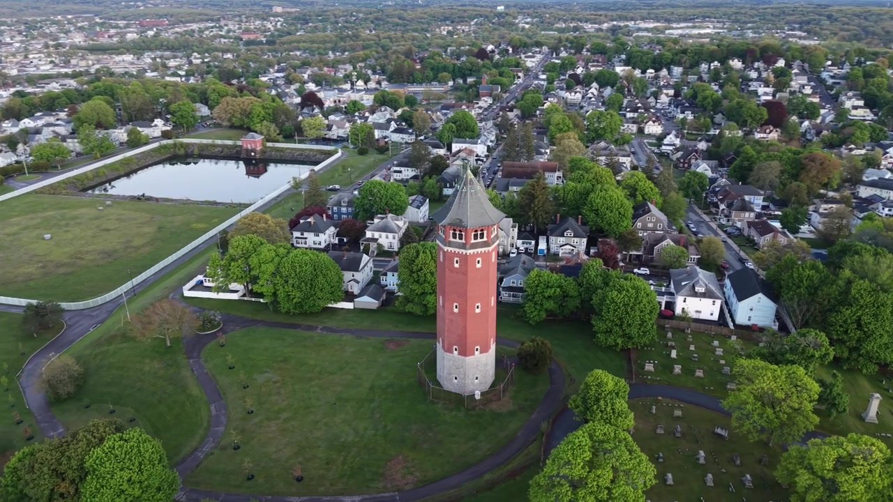 Histórica torre de agua de ladrillo rojo emergiendo de un parque verde con estanque reflectante, bloques residenciales y un horizonte urbano con campanarios de iglesias que se extienden hasta el horizonte. Massachusetts, EE. UU. Toma aérea orbital.