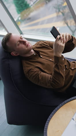 man rests in a soft chair with a phone. young man sitting near the window with a city transport background and using his mobile phone. cup of coffee on small table. top view. vertical video