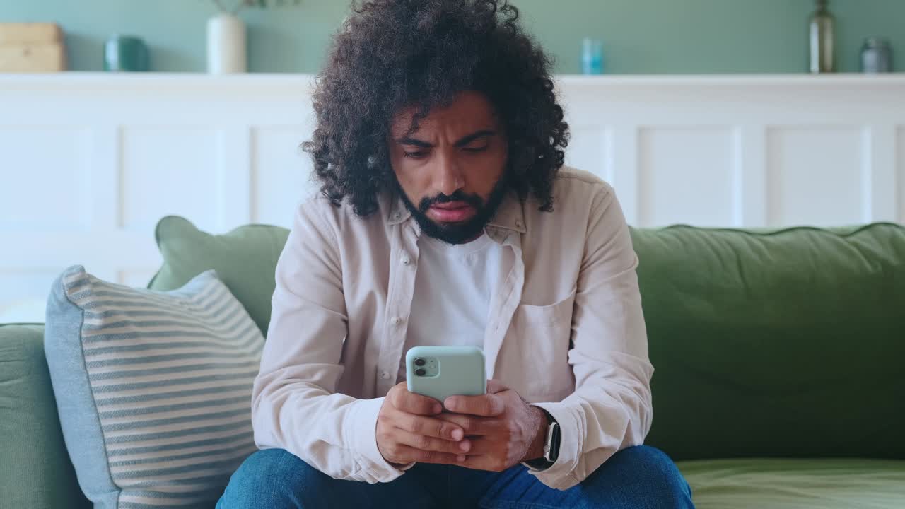 Young disgruntled arabian man nervously drops smart phone sits on sofa