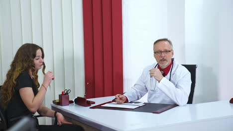 professional medic sits at table and speaks to a patient. senior doctor in glasses and grey beard. phonendoscope on the neck of a phisician.