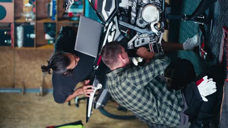 Vertical-video:-2-mechanics-repairing-a-bike.-A-brunette-man-in-a-gray-T-shirt-looks-at-the-screen-of-a-gray-laptop-and-a-man-in-a-checkered-shirt-repairs-a-motorcycle-in-a-workshop-garage