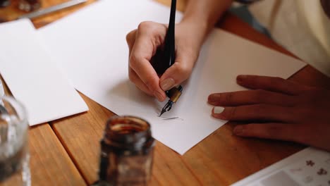 Woman-is-doing-calligraphy-with-an-ink-pen-on-paper,-styling-out-letters,-making-written-layout