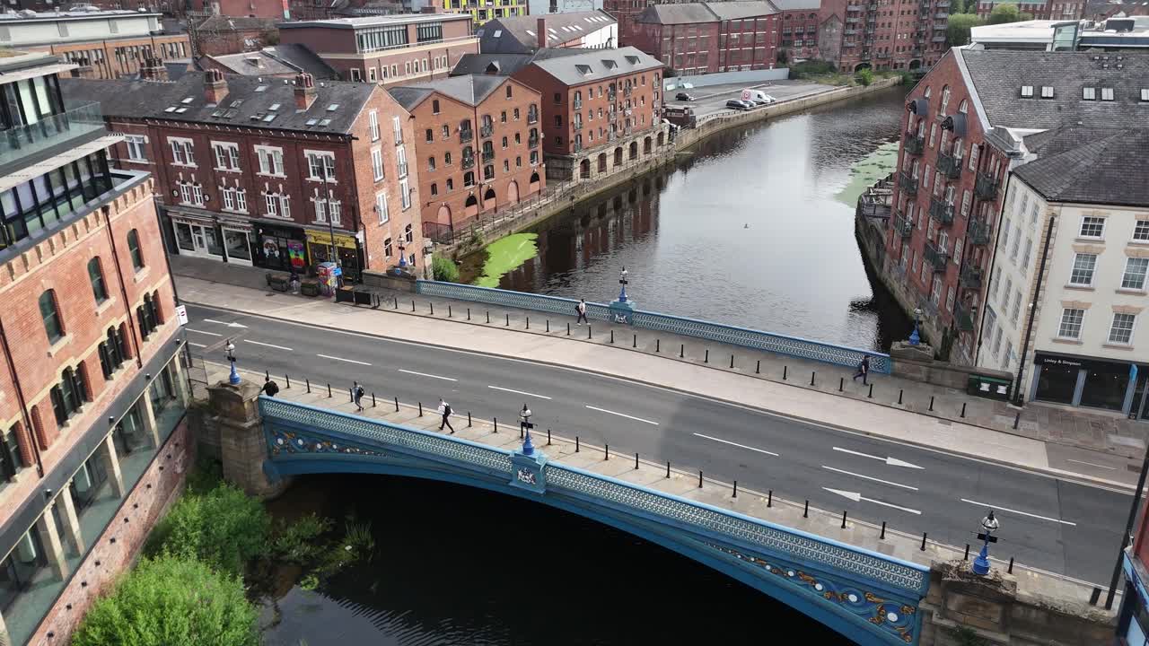 Aerial view of a green double-decker bus driving across a Victorian bridge above the River Aire, surrounded by historic city buildings under daylight