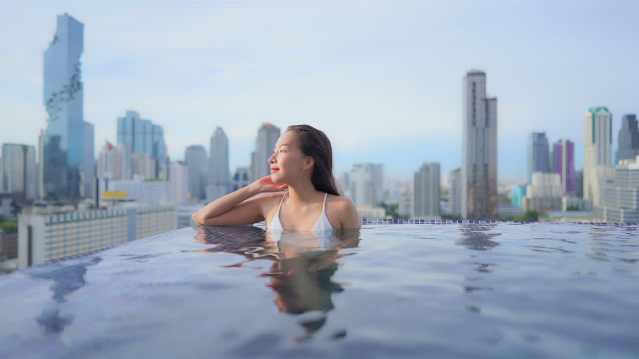 mujer joven en traje de baño blanco dentro de la piscina infinita de la azotea en el increíble fondo borroso del paisaje urbano de bangkok