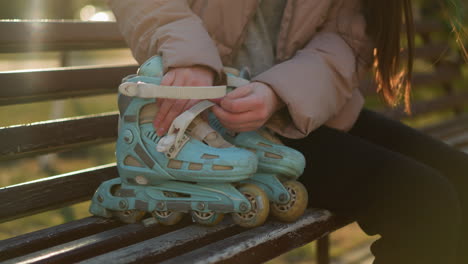 a girl wearing a peach jacket is seated on a park bench, preparing to put on her blue rollerblades. the close-up shot captures her hands as she adjusts the straps, readying herself for the activity