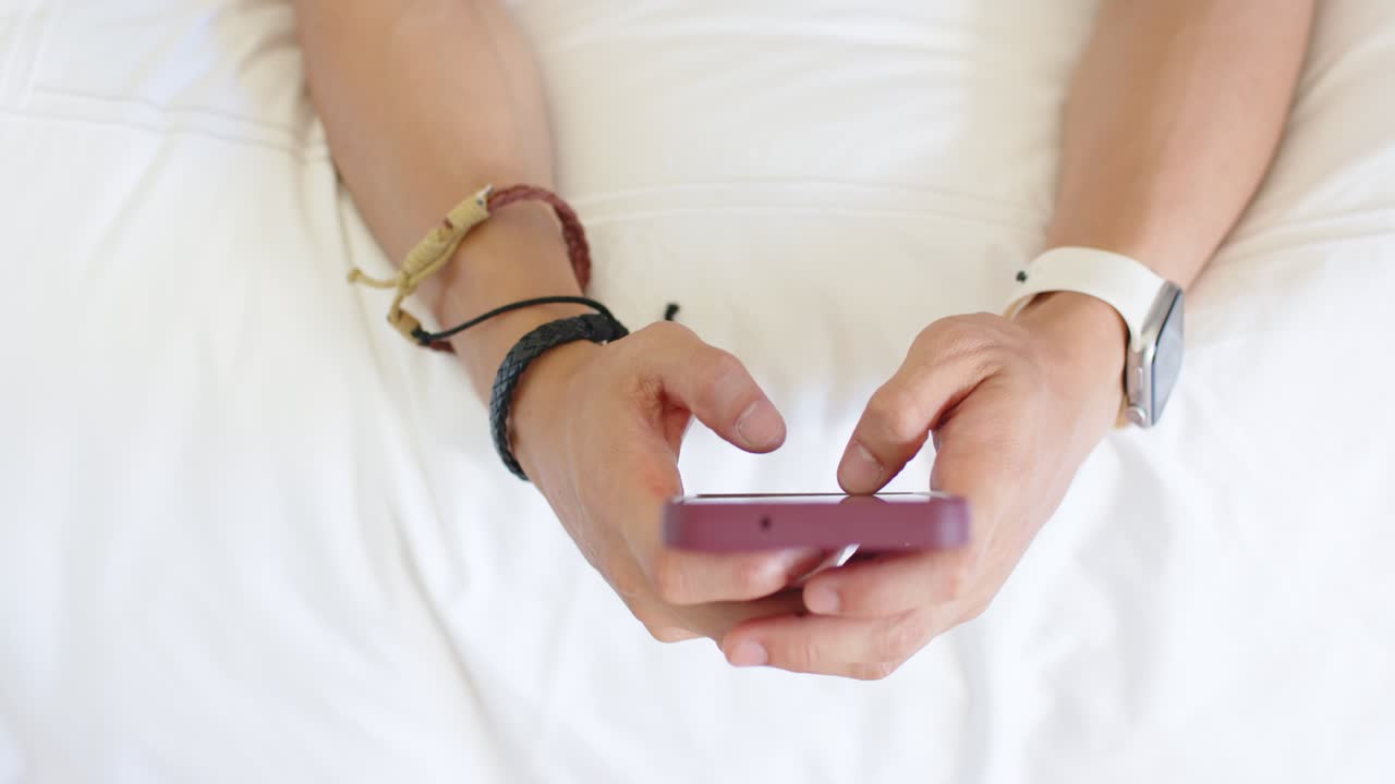 Overhead hands are holding purple case phone, tapping, swiping on white bed, bracelets and watch