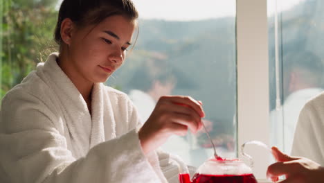 lady stirs tea in glass teapot with spoon closeup. young woman prepares natural hibiscus tea for boyfriend. man holds hot teapot helping girlfriend