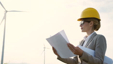 Caucasian-woman-engineer-wearing-a-helmet-watching-some-blueprints-at-wind-station-of-renewable-energy