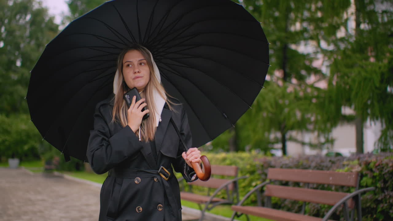 Beautiful female wearing black coat and hood holding umbrella and talking on phone while walking near bench in park on cloudy rainy day, creating urban lifestyle communication scene with greenery background