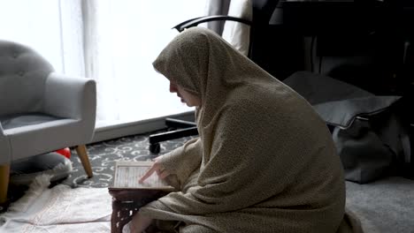 seated on the floor and covered with a pile of clothing, a young girl reads a religious book, reflecting the idea of studying and learning within the comfort of home