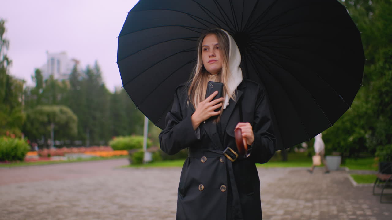 검은색 코트와 후드를 입고 우산을 든 여성이 흐리고 비 오는 날 공원에서 통화하며 걷고 있고, 주변은 초록빛 식물과 흐릿한 꽃들로 둘러싸인 도시적인 라이프스타일의 소통 분위기