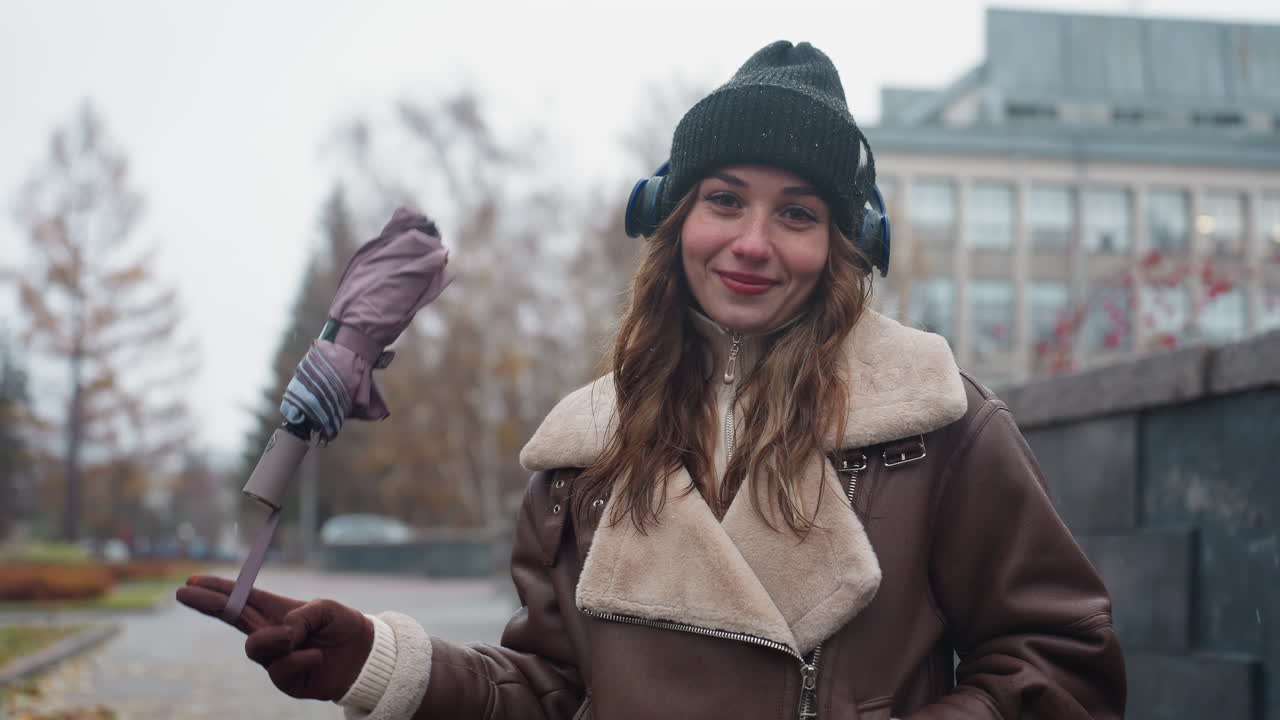 Portrait view of cheerful woman in black knit cap brown shearling jacket smiling while standing outdoors on concrete path with autumn leaves swinging umbrella playfully during chilly day in soft light