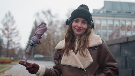 portrait view of cheerful woman in black knit cap brown shearling jacket smiling while standing outdoors on concrete path with autumn leaves swinging umbrella playfully during chilly day in soft light