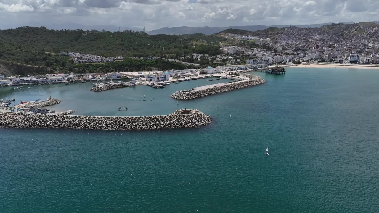 Ampla vista aérea de um porto mediterrâneo no Norte de Marrocos, mostrando pequenos veleiros, múltiplos quebra-mares protetores, água azul calma e colinas costeiras verdes cercando a marina