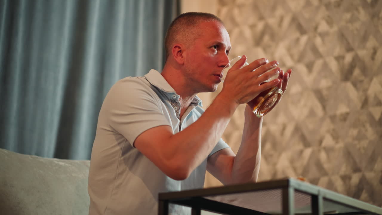 Man eating meal lifts glass of juice to drink and drops it back with satisfied look while sitting indoors, textured wall background, relaxed mood, enjoying refreshing beverage after food