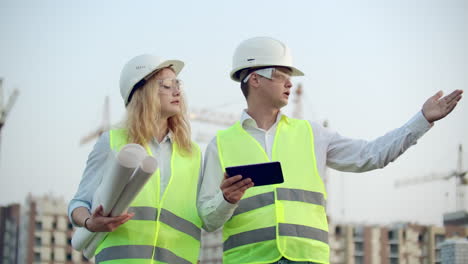 Zwei-Wirtschaftsingenieure-Tragen-Schutzhelme-Und-Halten-Ein-Tablet-In-Der-Hand.-Sie-Arbeiten-Als-Ingenieure-Und-Besprechen-Die-Inspektion-Von-Zeichnungen.-Draußen-An-Einem-Gebäude.-Technische-Werkzeuge.