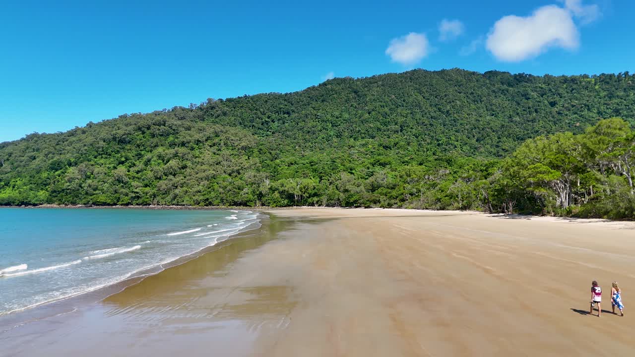 A couple strolls along a secluded beach in Port Douglas, under clear skies with lush greenery and gentle waves