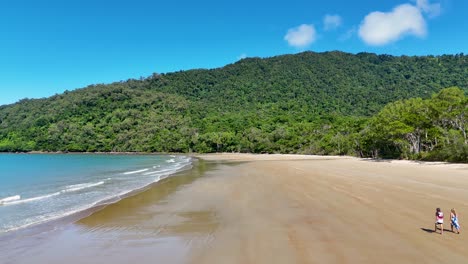 A couple strolls along a secluded beach in Port Douglas, under clear skies with lush greenery and gentle waves
