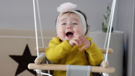 지나치게 흥분한 아기가 집에서 도어케이스 스윙에 앉아 박수를 치고 손을 움직이며 웃고 있습니다.