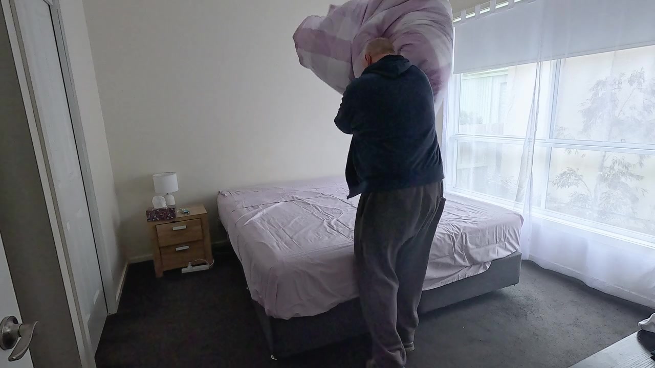 Uomo di mezza età si innervosisce mentre cerca di mettere un copripiumino su un piumone in una camera da letto modesta