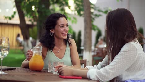 Concepto-De-Personas,-Comunicación-Y-Amistad-Mujeres-Jóvenes-Sonrientes-Bebiendo-Jugo-De-Naranja-Y-Hablando-En-Un-Café-Al-Aire-Libre-En-Verano