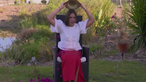 A young girl sits in a garden chair and tips a straw hat with flower petals over her head