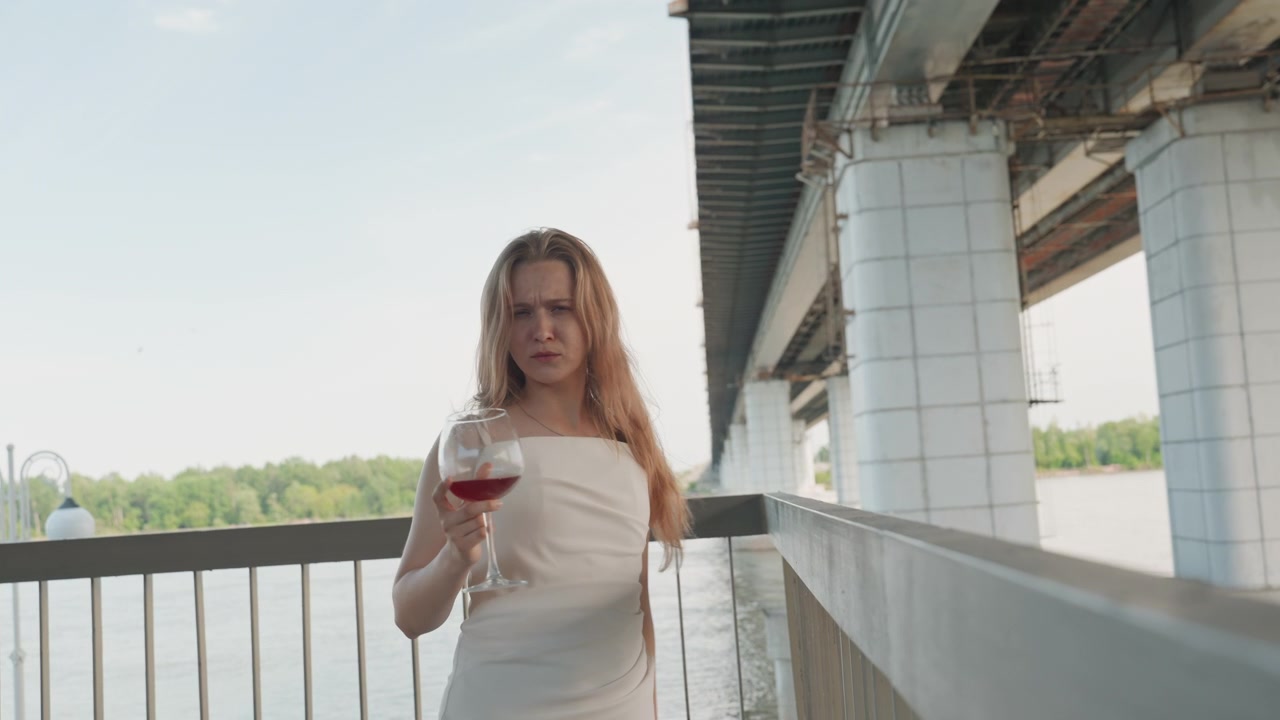 Donna in abito bianco cammina lungo un ponte esterno, tenendo e bevendo da un bicchiere di vino rosso. La scena presenta elementi architettonici come pilastri di cemento sotto la struttura con alberi verdi.
