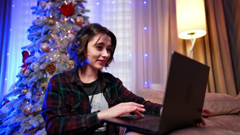 dark-haired girl in checkered shirt opens her laptop. woman works on computer online at christmas.