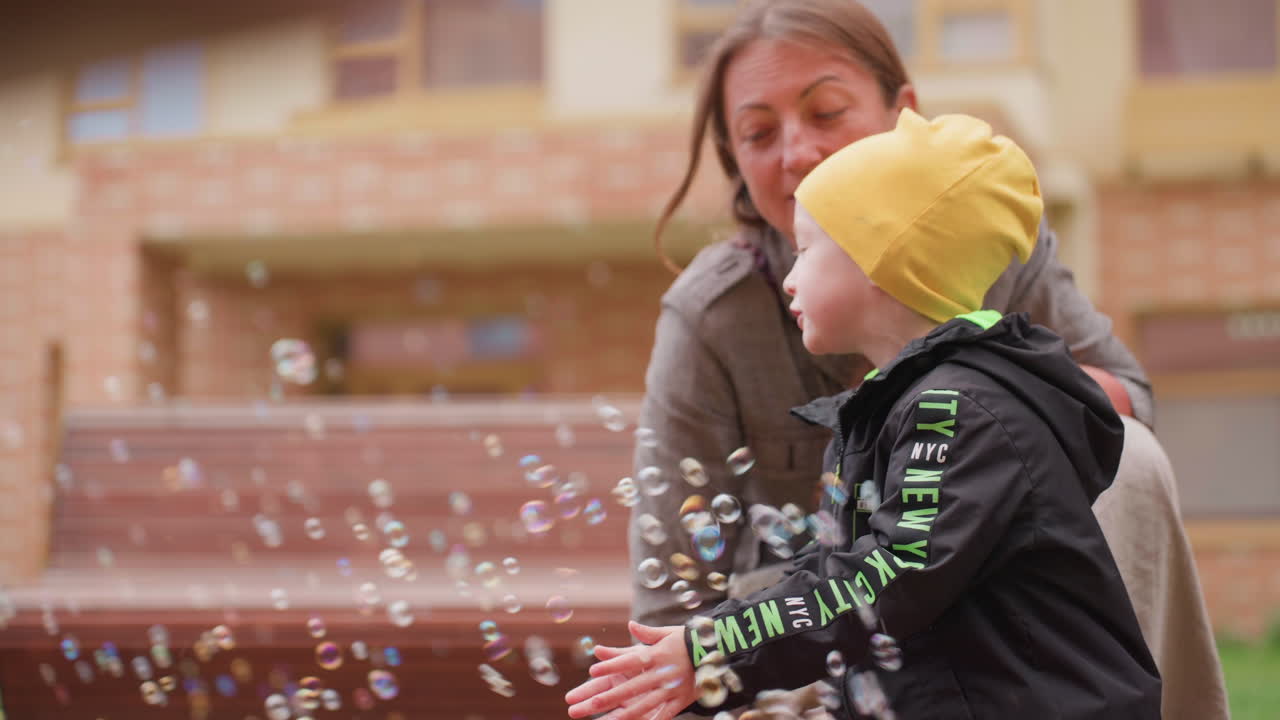 Mother leans close speaking to son as child reaches to catch floating soap bubbles, soft blur benches and windows behind, gentle guidance, warm courtyard moment showing curiosity