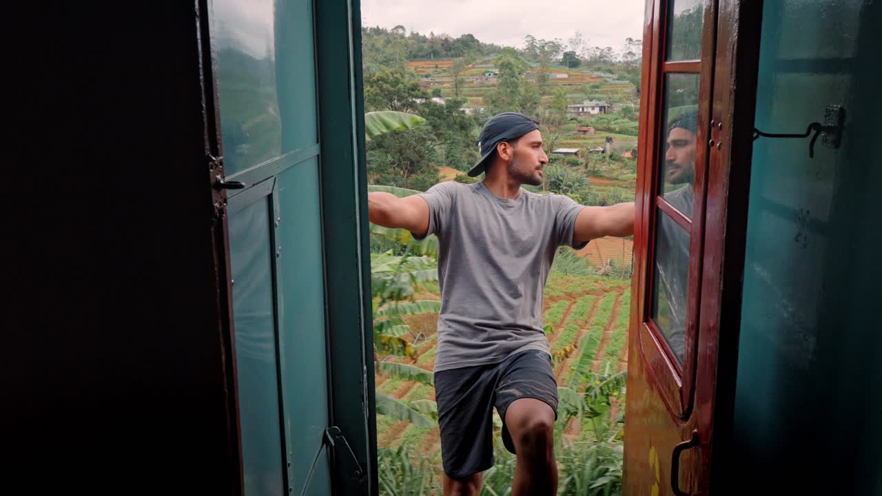 A man rides with the door open on the scenic train route from Ella to Kandy in Sri Lanka. Surrounded by lush greenery and tea plantations.
