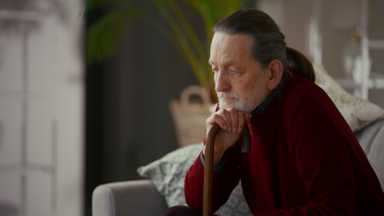 Thoughtful Elderly Man Sitting with Walking Stick at Home