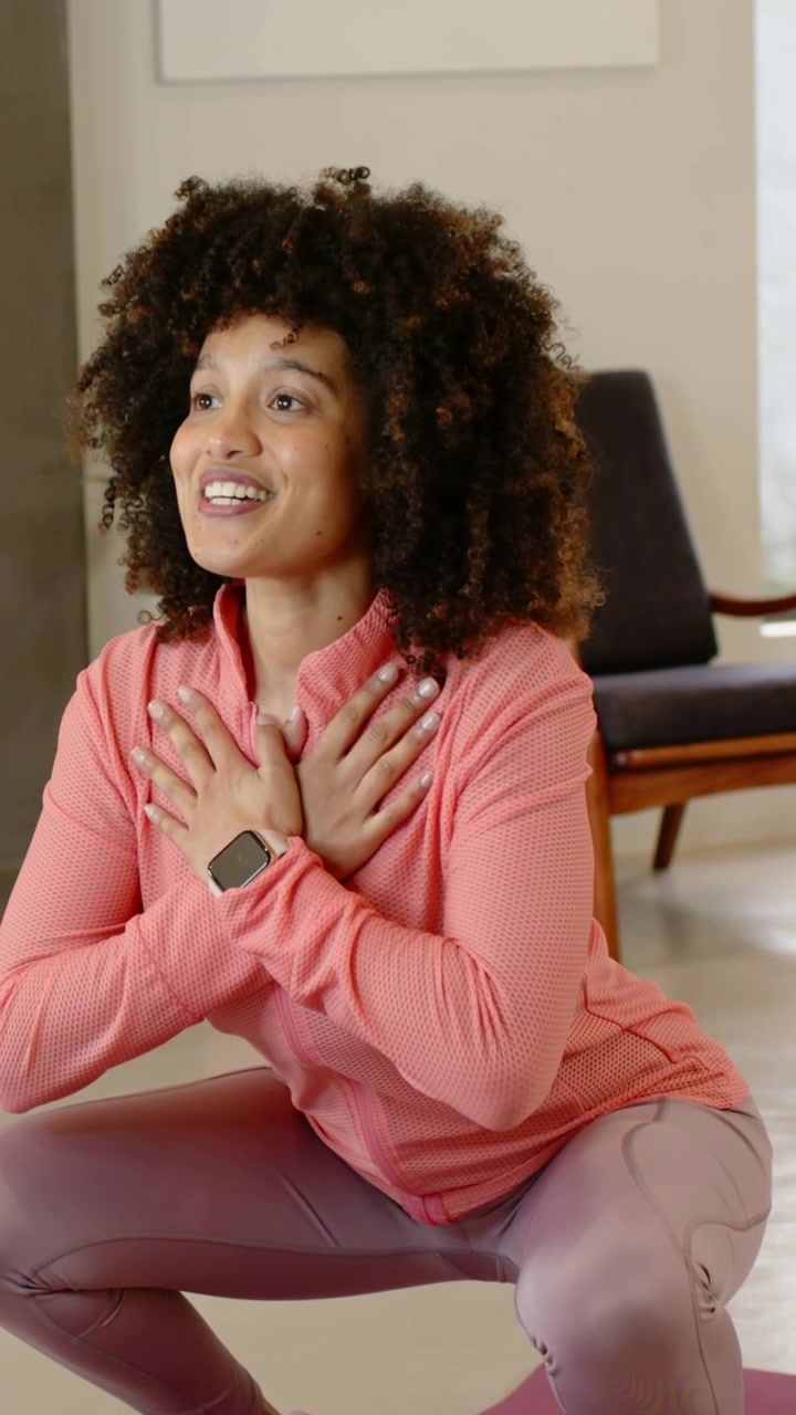 Vídeo vertical: Mulher de jaqueta coral começando, levantando de um agachamento em tapete roxo em casa com smartwatch