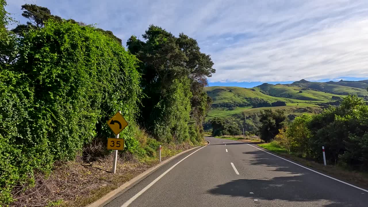 Forward-facing camera captures a winding rural road bordered by dense greenery and rolling hills under bright daylight, highlighting natural landscapes and smooth motion