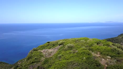 drone aerial shot, flying over a cliff covered in green vegetation towards a wonderful blue mediterranean sea along the coast of sardinia in italy