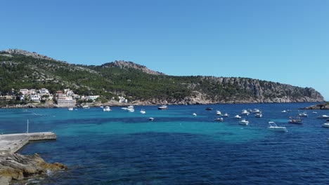 drone flying over crystal clear blue waters in a quiet natural harbour with boots and yachts coming into sight and mallorca landscape in the background