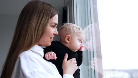 Long-haired cheerful woman holding a blond baby. Mother with child stands at the window pointing at something.