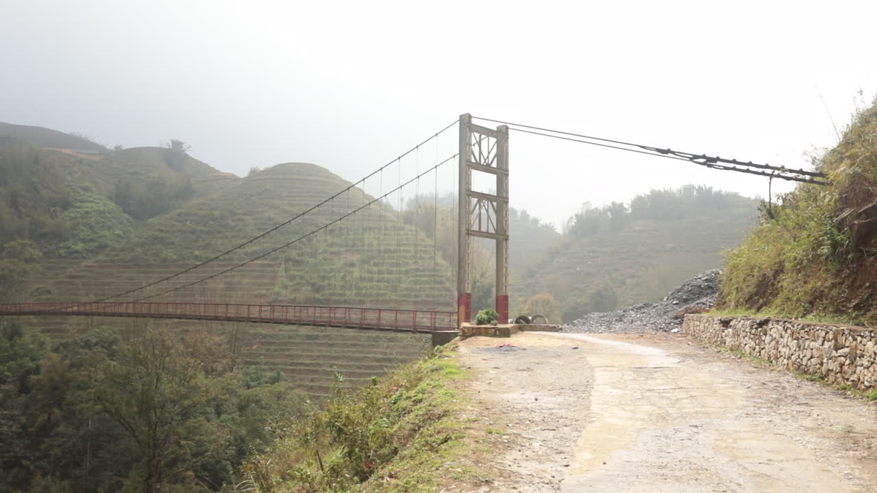 Suspension bridge over misty hills in rural Vietnam with a dirt road leading to it, creating a tranquil scene