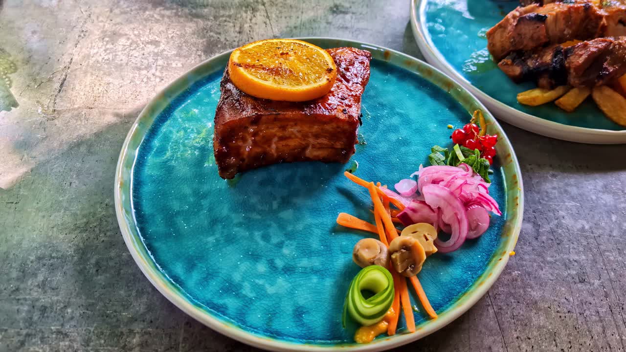 Close-up of a plated restaurant dish featuring glazed meat with an orange slice, served with colorful pickled vegetables and garnishes on a vibrant blue ceramic plate