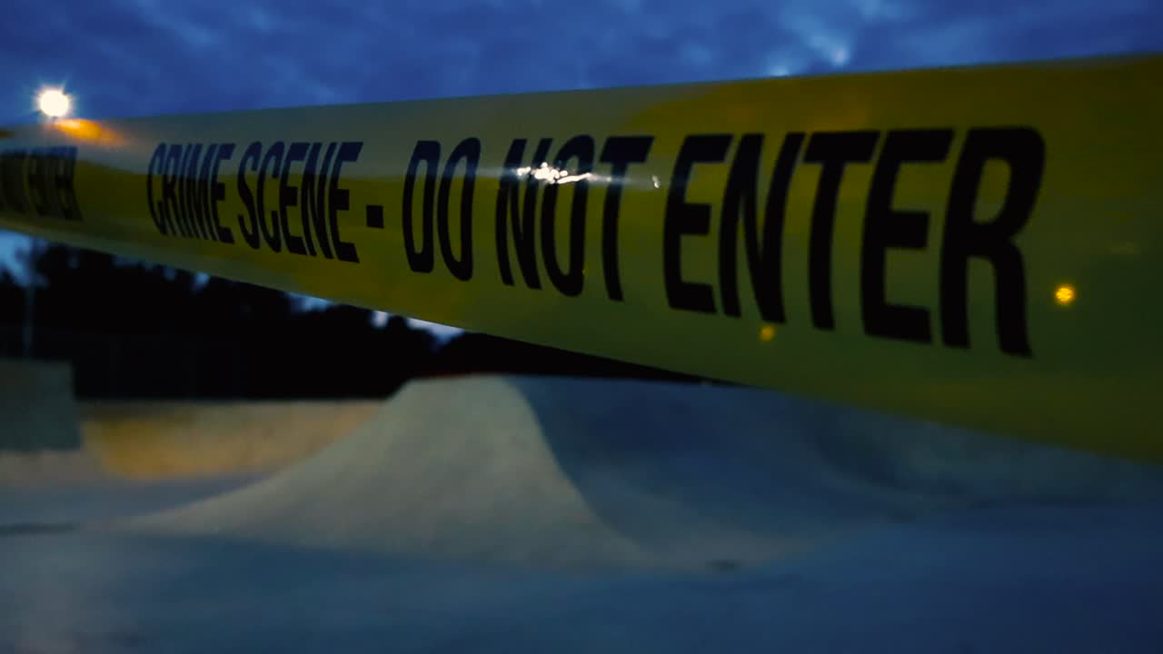 Slow tracking shot along crime scene tape fluttering in the wind in front of shadowy closed skatepark at evening, night. Police tape pulled across low light restricted park area with street lights