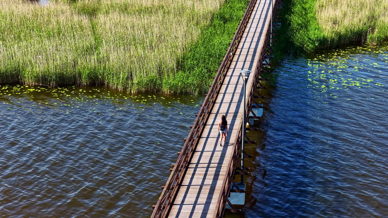 Woman walks alone on Širvėnos Lake Bridge surrounded by rippling water and reeds, aerial view