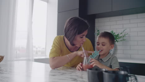 madre hermosa y hijo juntos sonriendo bebiendo jugo de naranja de un vaso de vidrio a través de un tubo